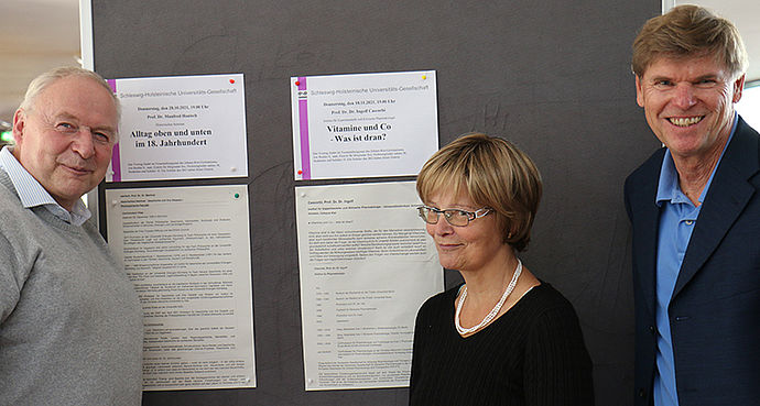 Zwei Männer und eine Frau vor einer Infotafel mit Werbeblättern für Veranstaltungen der Universitätsgesellschaft.