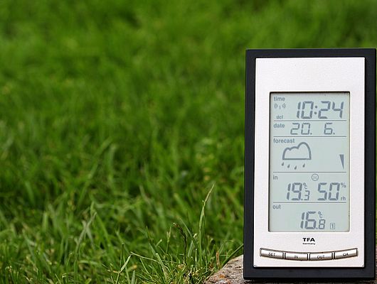 Eine digitale Wetteranzeige zeigt Werte. Dahinter sieht man grünen Rasen. 