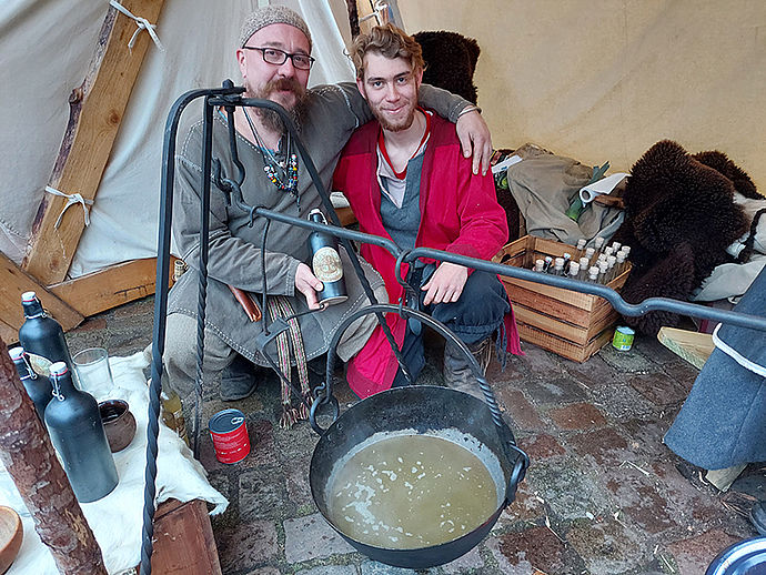Zwei Männer in mittelalterlicher Kleidung hinter einem dampfenden Kochtopf.