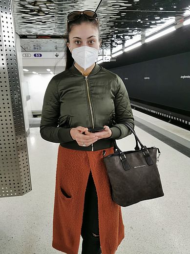 Junge Frau mit Maske, graue Jacke, Handtasche in einer S-Bahnstation