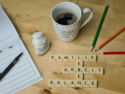 Eine Tasse Kaffee, Stifte und ein Terminkalender sind um Buchstabenbausteine angeordnet, die die Worte Familie, Arbeit, Leben und Balance formen 