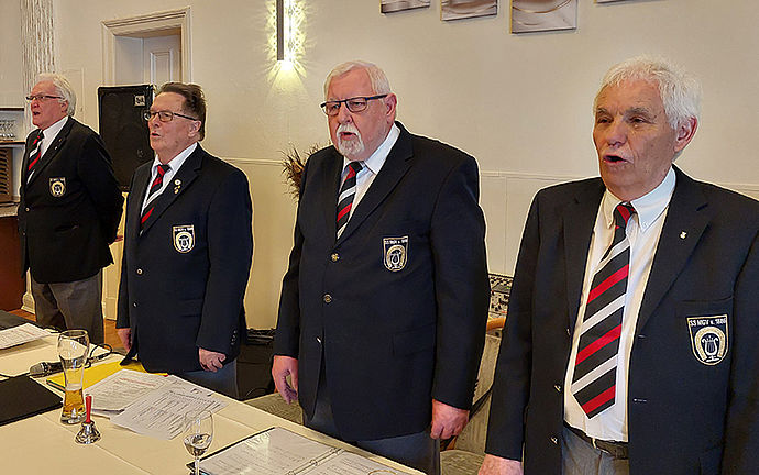 Vier Männer stehen offensichtlich fröhlich singend nebeneinander hinter ihren Tischen.