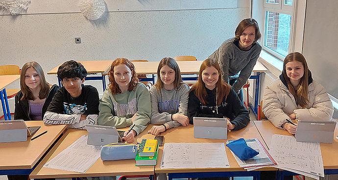 Fünf Schülerinnen, ein Schüler und eine Lehrerin lächeln hinter ihren Tablets in die Kamera