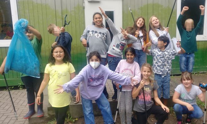 13 Kinder, drei davon gepixelt, posieren mit Gartengeräten zum Müllsammeln.